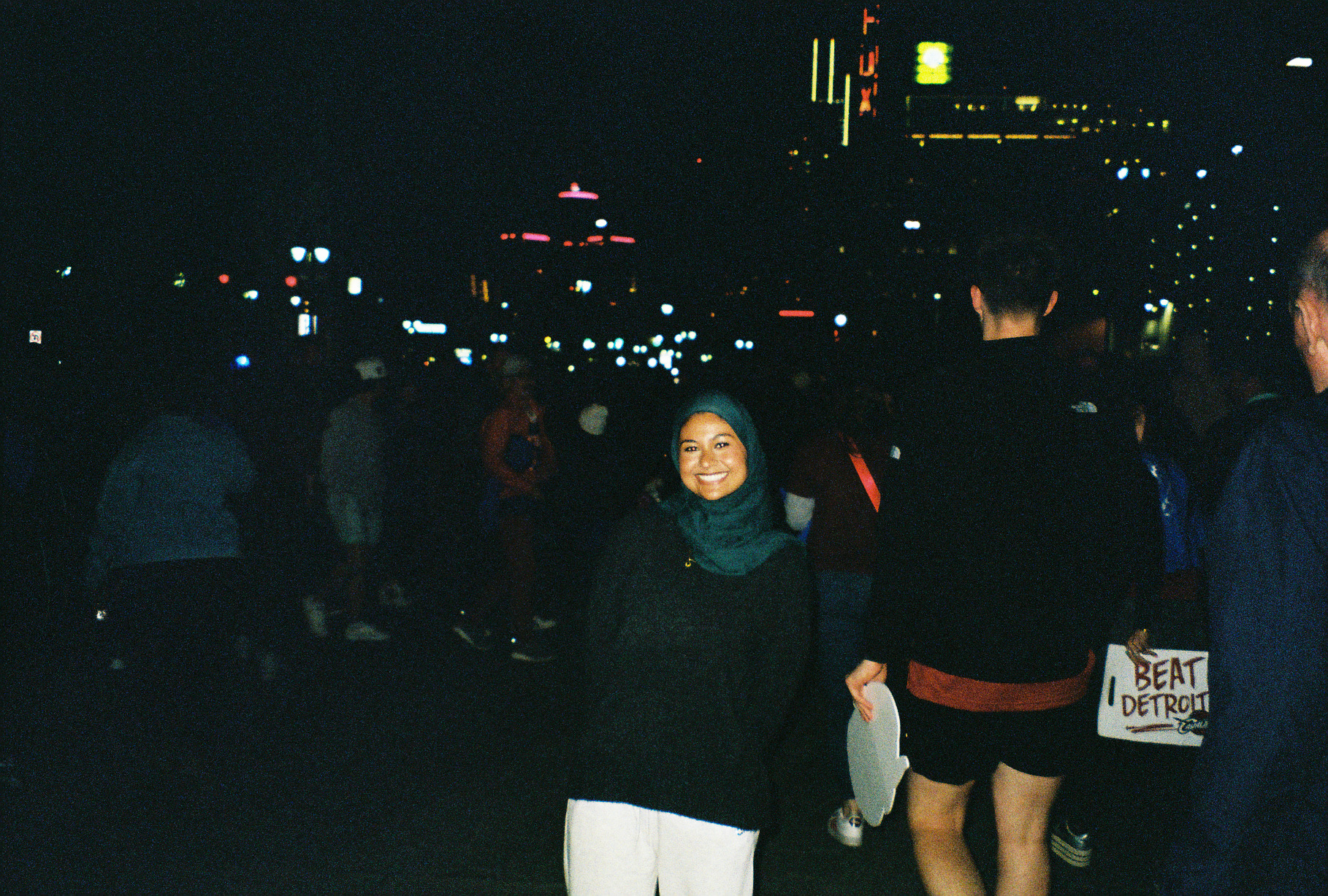 Tabassum smiling on a lit-up city street at night, a "Beat Detroit" sign held behind her
