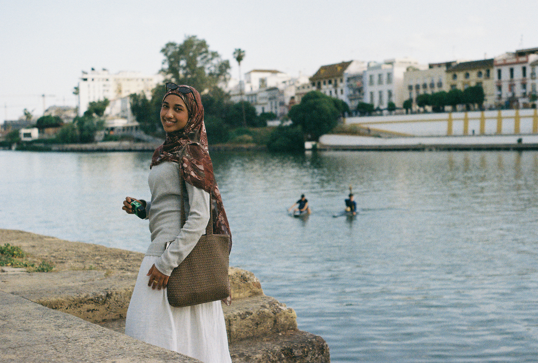 Tabassum looking back at the camera beside a river in Seville, two people paddling small boats behind her