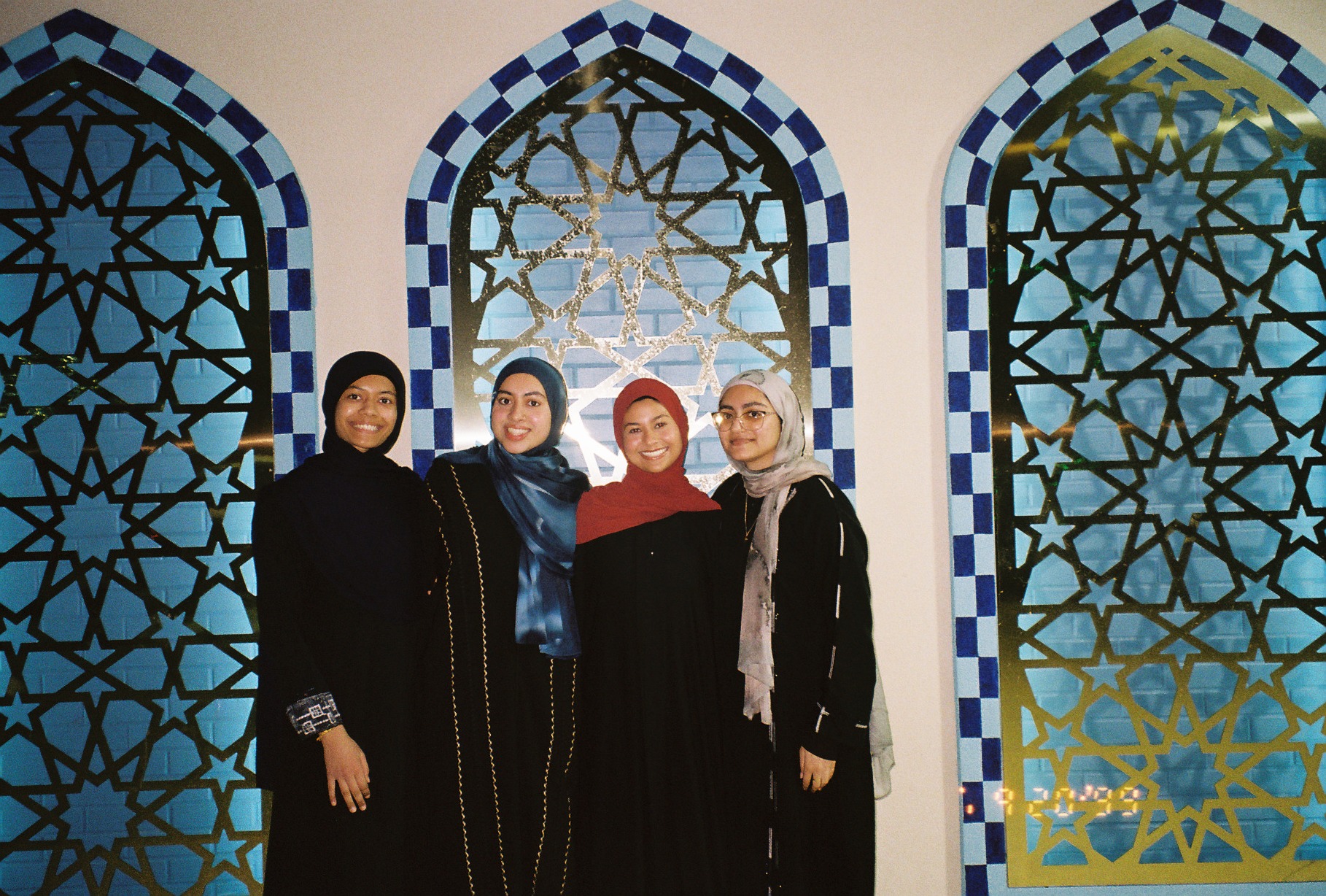 Four young women in hijabs and abayas standing together smiling in front of a mosque