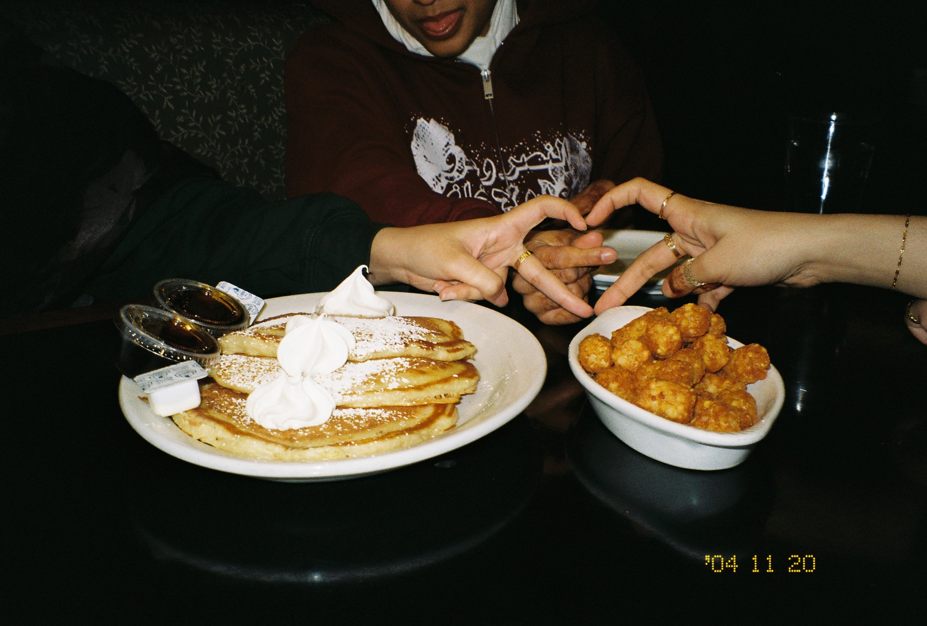 Pancakes and tater tots on a dark table at Pizza House, two sets of hands making a heart above the food