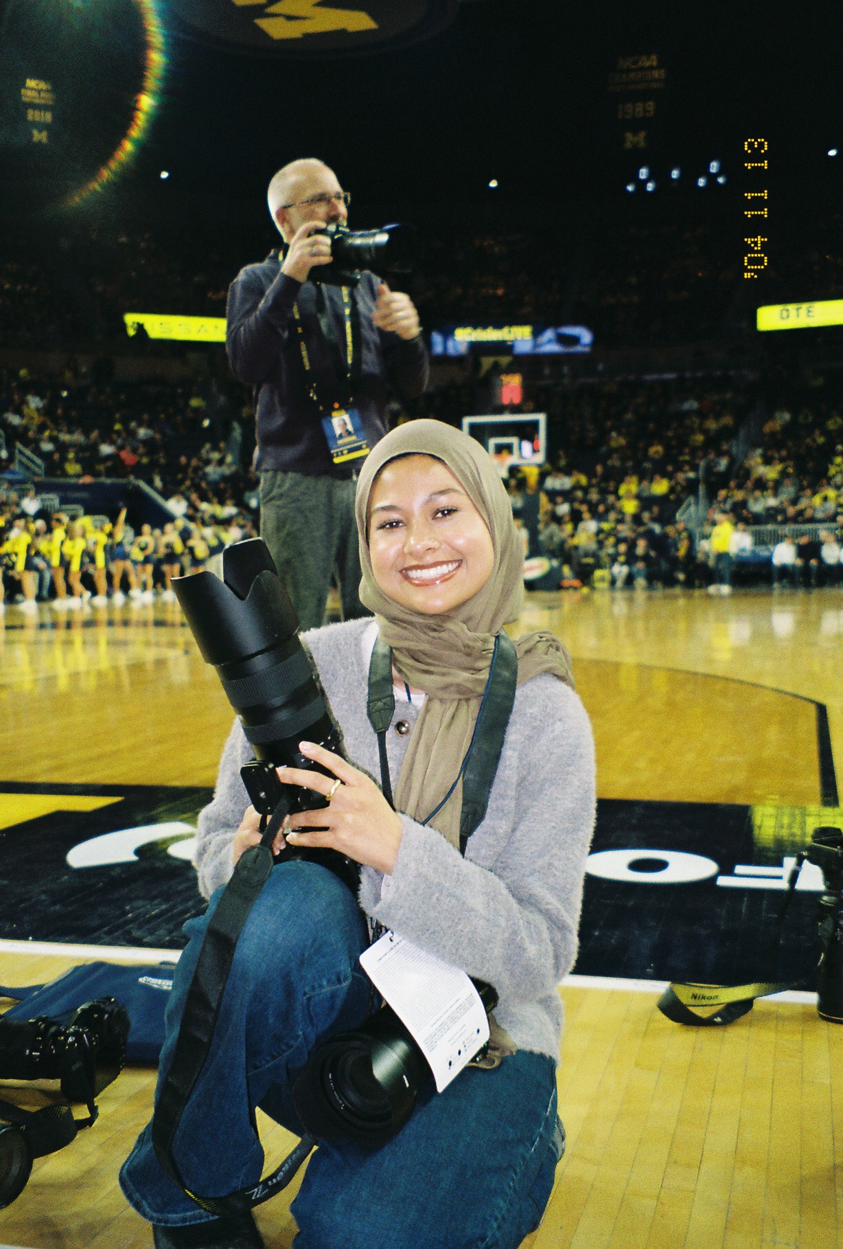 Tabassum kneeling on the Crisler Arena court holding a camera, smiling