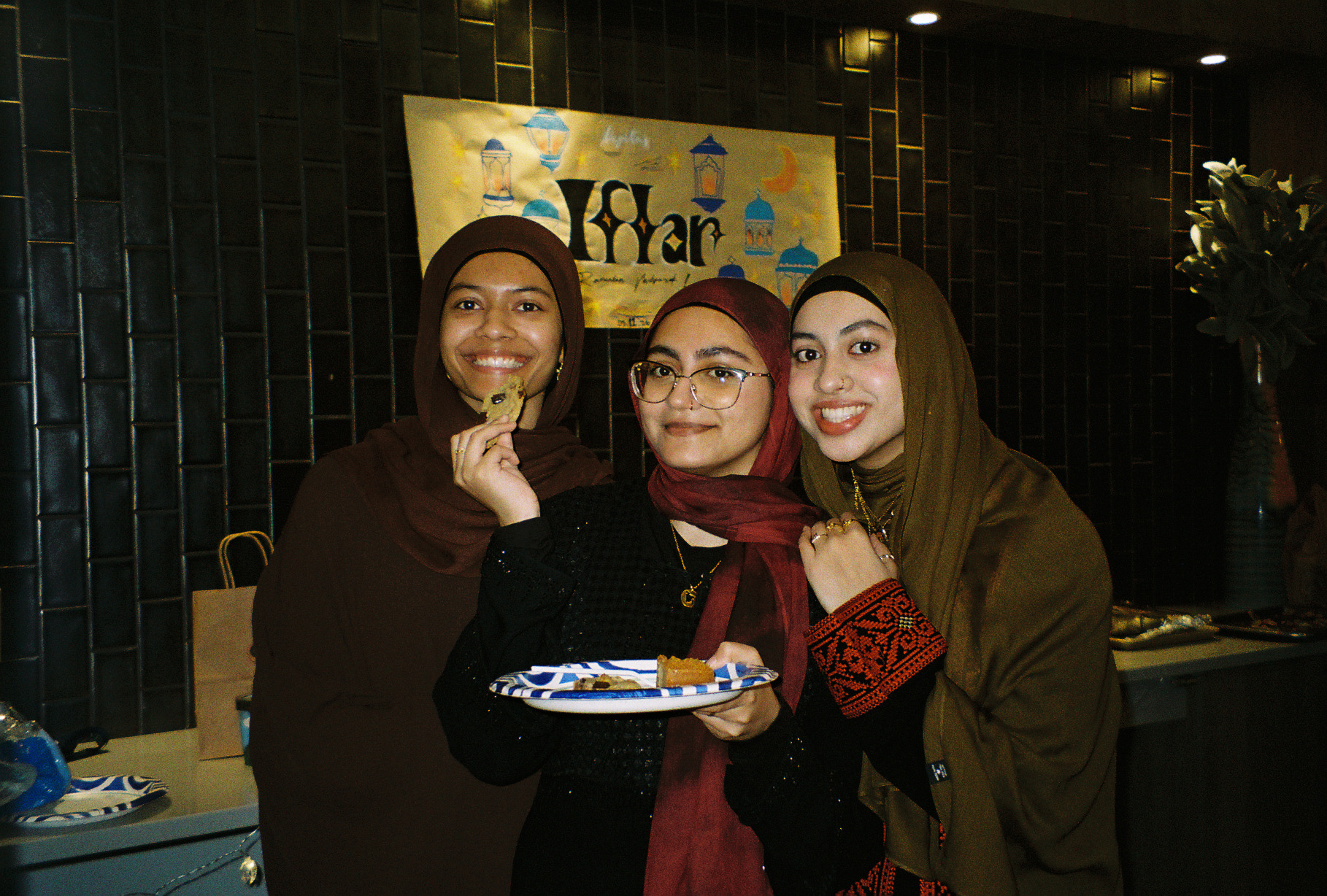 Three friends at an Iftar event in front of a hand-painted Ramadan banner, one holding a plate of food